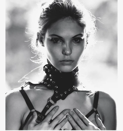 Monochrome portrait of a young woman with fair skin, hands raised to her chest, wearing a studded leather harness and choker.