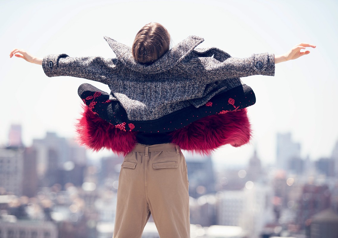 Woman in coat stands on rooftop with arms outstretched, city skyline in background, creating a fashion editorial image with outdoor setting.