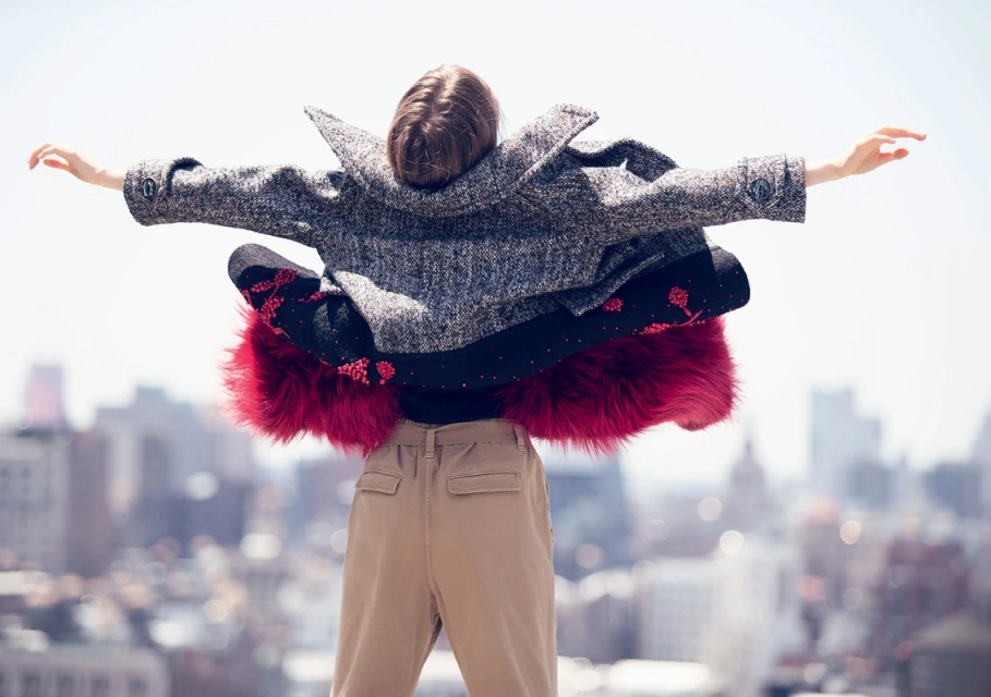 Woman in coat stands on rooftop with arms outstretched, city skyline in background, creating a fashion editorial image with outdoor setting.