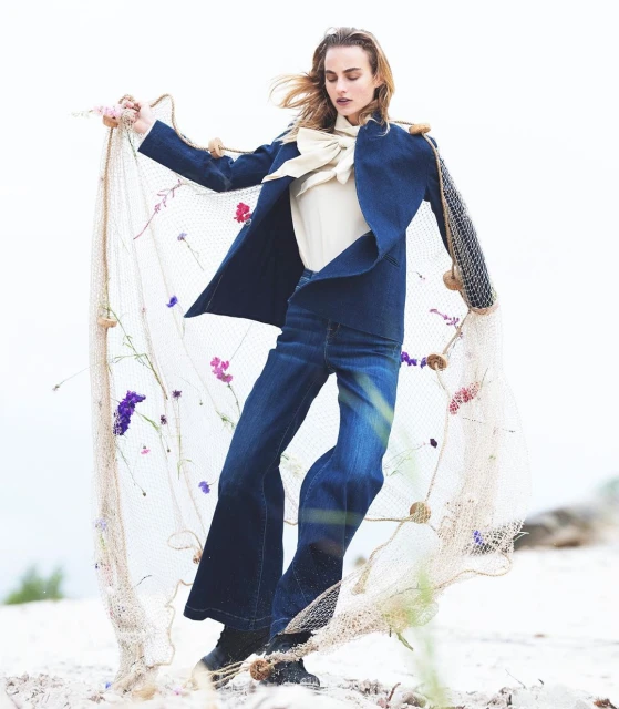 Fashion shot of a woman in a blue blazer and jeans holding a fishing net with flowers on a light sandy surface.