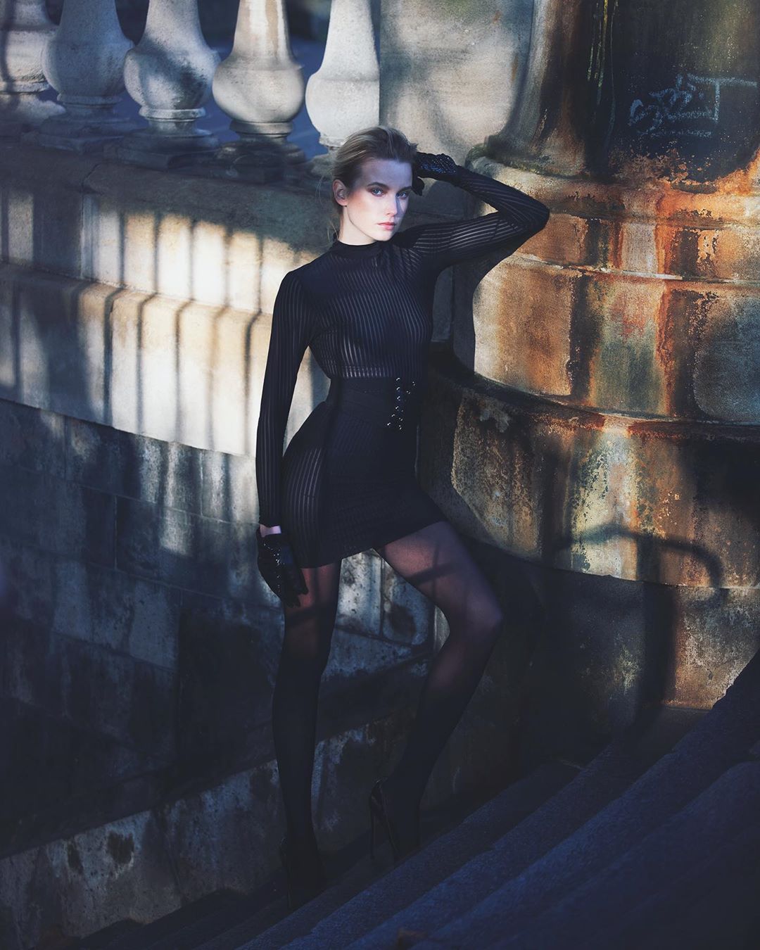 Woman in black dress and tights poses on stone steps near a balustrade, with natural light and shadows creating a striking fashion image.