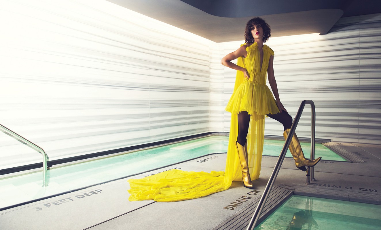 A fashionable young woman in a yellow dress and boots poses at the edge of a pool in a sleek indoor setting, showcasing an editorial style.