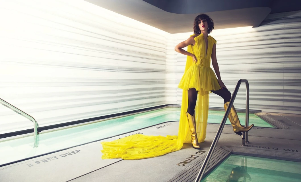 A fashionable young woman in a yellow dress and boots poses at the edge of a pool in a sleek indoor setting, showcasing an editorial style.