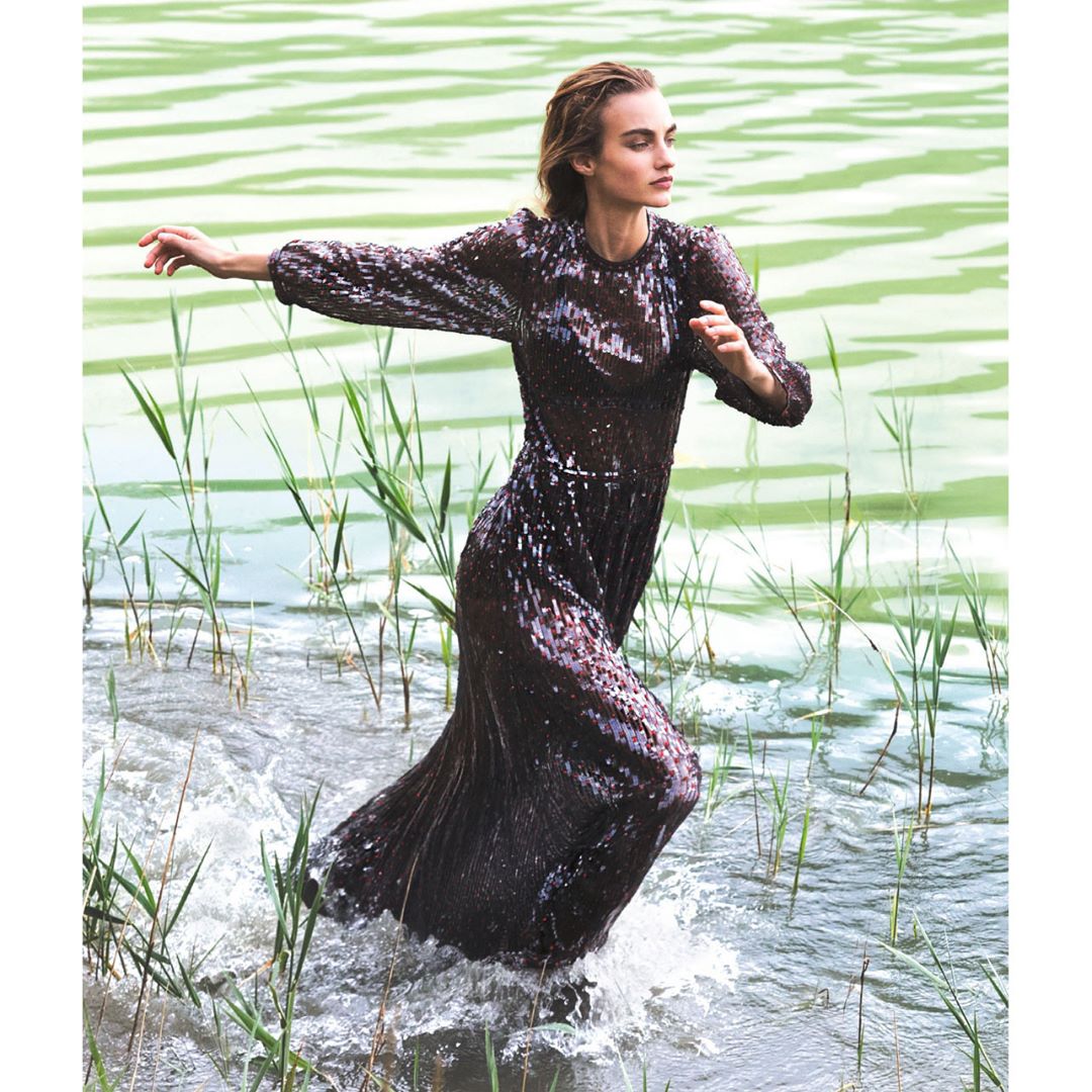 A slender woman in a dark sequin dress stands in a lake with green water and tall reeds, arms outstretched, making splashes in the water.
