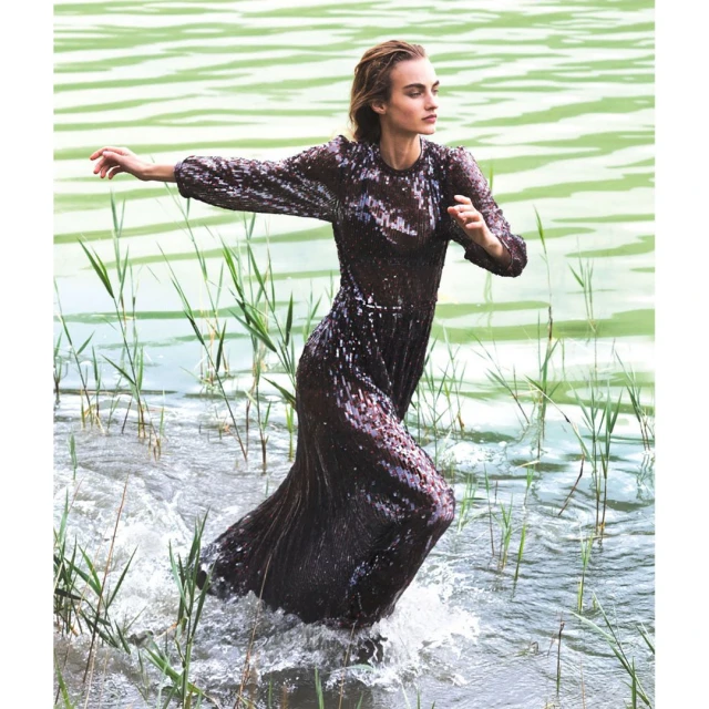 A slender woman in a dark sequin dress stands in a lake with green water and tall reeds, arms outstretched, making splashes in the water.