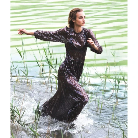 A slender woman in a dark sequin dress stands in a lake with green water and tall reeds, arms outstretched, making splashes in the water.
