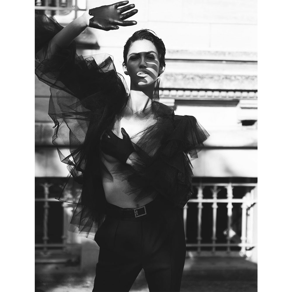 Monochrome fashion shot of a model in dark tulle top, gloves, and pants with a belt, posing near a building with natural shadows.