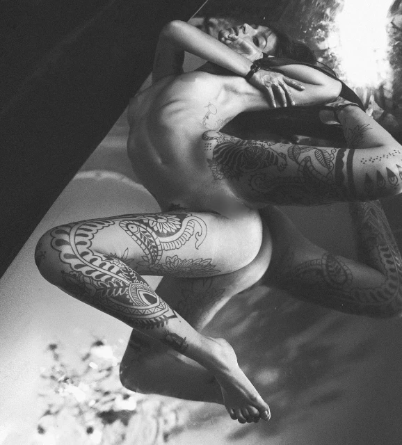 Black and white high-angle view of a tattooed adult embracing another person while lying on a reflective surface in an indoor setting.