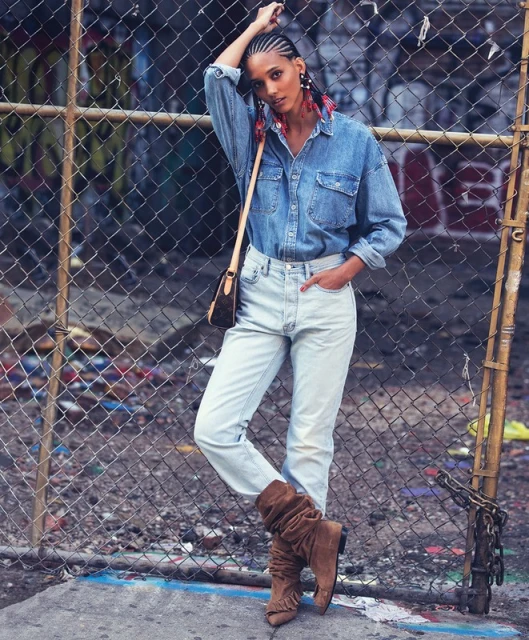 Woman in denim shirt and jeans poses against a chain-link fence in an urban setting, showcasing fashion and urban style.
