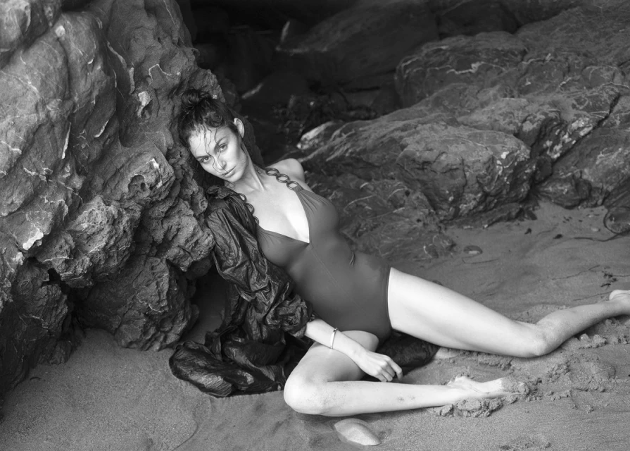 Monochrome portrait of a woman in a swimsuit reclining on a sandy beach with rock formations, creating a dramatic and striking visual.