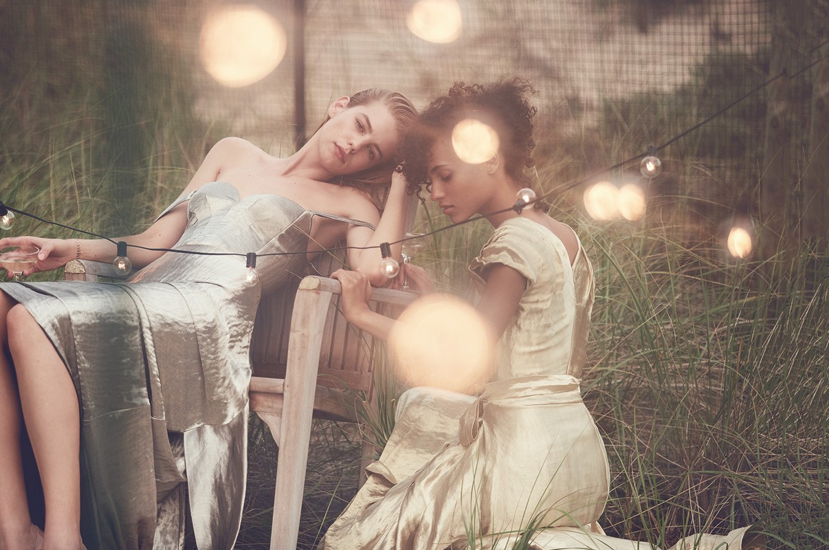 Two women in elegant gowns sit on a wooden chair amidst tall grass, illuminated by string lights, creating a whimsical outdoor scene.