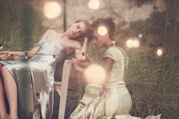 Two women in elegant gowns sit on a wooden chair amidst tall grass, illuminated by string lights, creating a whimsical outdoor scene.