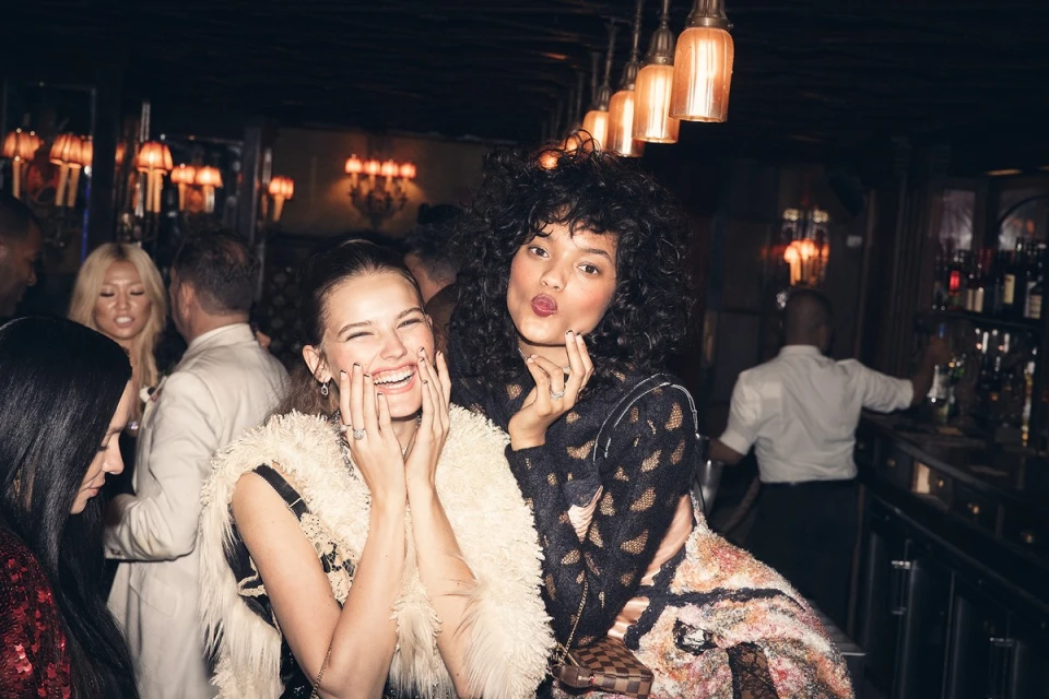 Two young women pose in a warmly lit bar, one smiling with hands on cheeks, the other with curly hair making a kiss face.