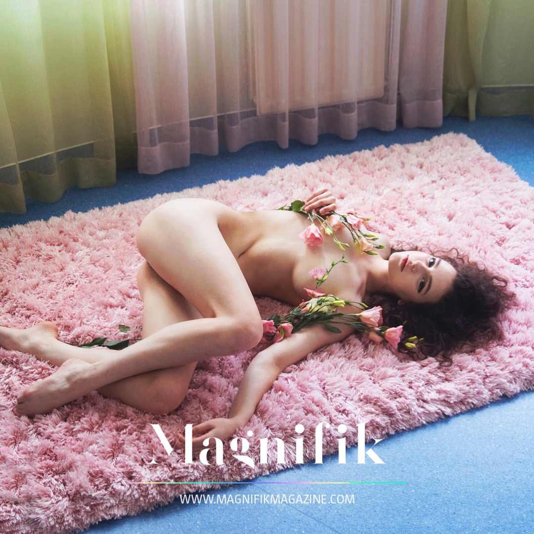 Nude female fashion model poses on pink shag rug with rose branches in indoor photo studio with soft, diffused lighting.