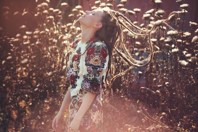 Woman with dreadlocks stands in a field of wildflowers, wearing a floral dress and gazing upwards in a sun-drenched outdoor scene.