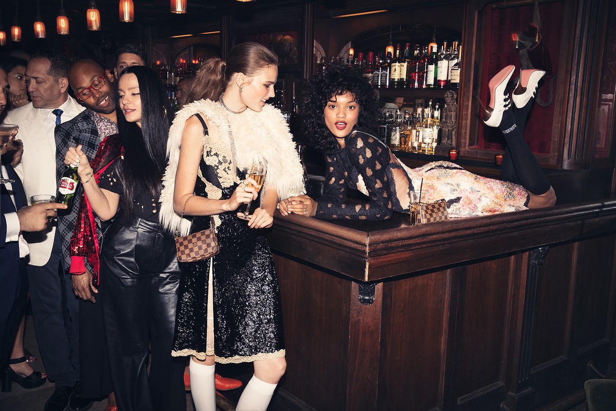 Two women pose at a bar with drinks and a group of people in the background, creating a vibrant, social atmosphere in an indoor setting.