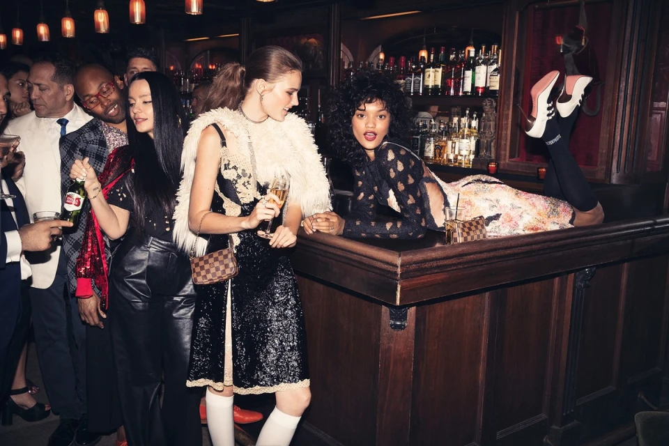 Two women pose at a bar with drinks and a group of people in the background, creating a vibrant, social atmosphere in an indoor setting.