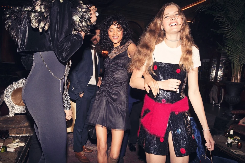 A group of young adult women at an indoor party wearing dresses and elegant attire in a dimly lit indoor setting with additional people.