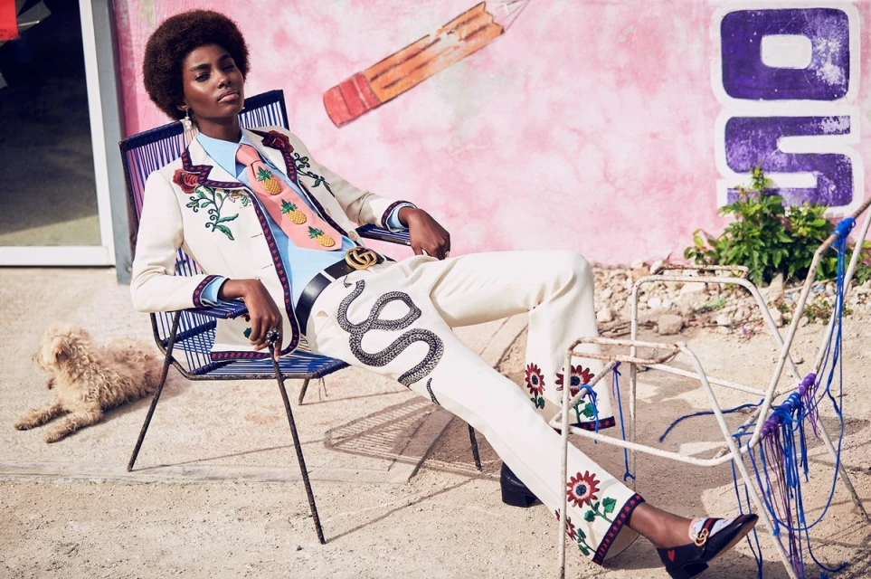 A stylish Black woman in a cream suit with snake embroidery poses in a chair with a small dog; a pink wall with graffiti is behind her.