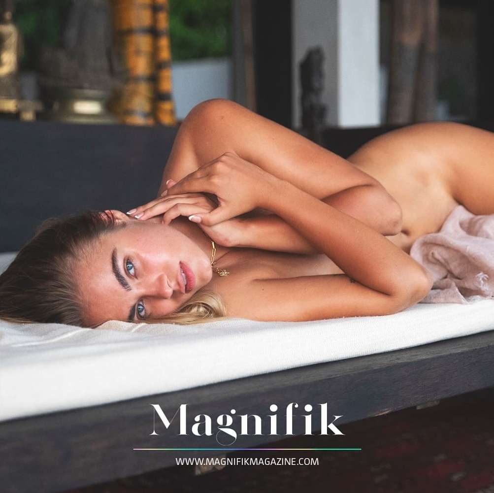 Blonde woman poses nude in an indoor setting on a white bed while looking at the camera. Her hands are near her face.