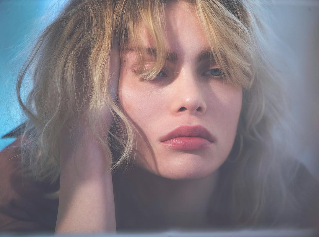 Close-up portrait of a young blonde woman with fair skin, gazing slightly upwards in a soft, diffused light setting. Facial expression is neutral.