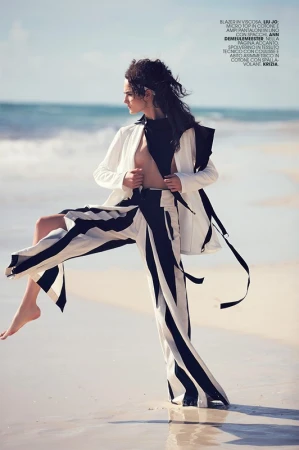 Model in wide striped pants and open white jacket posing dynamically on a sandy beach by the ocean.