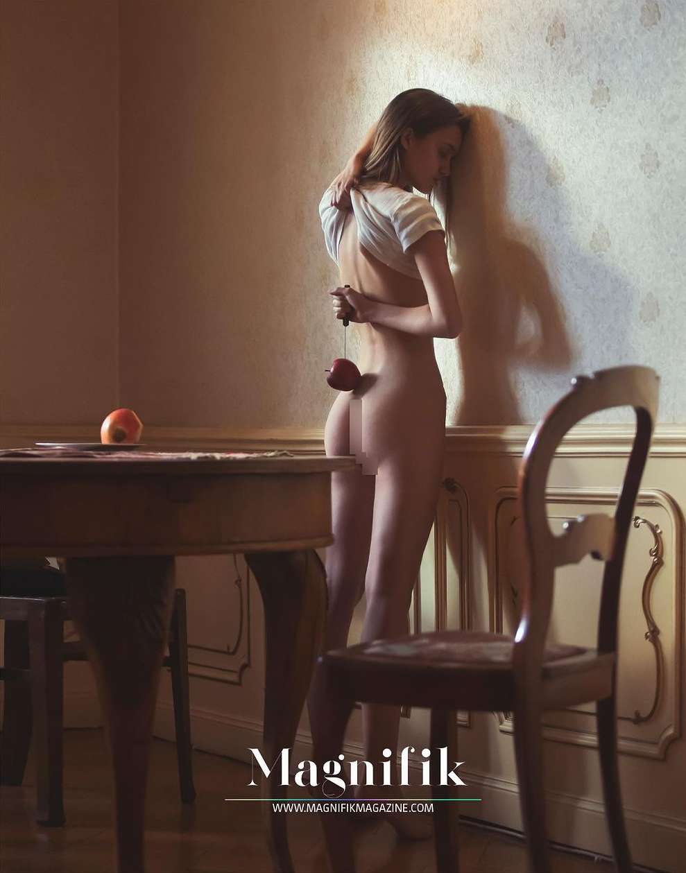 Artistic nude photo of a young adult woman posing with an apple tied behind her back in a warm-toned interior room setting.