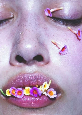 Close-up of a person's face with eyes closed and flowers on their lips and eyelids in a beauty or wellness concept image.