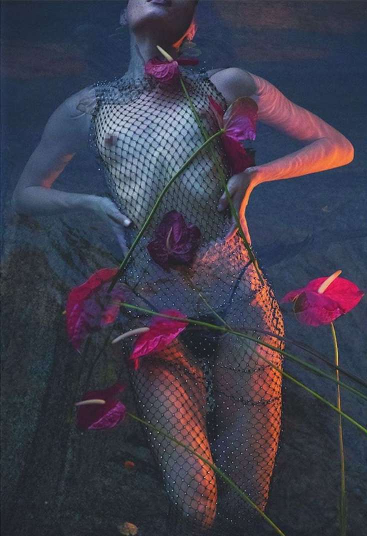 An adult woman with a net-like garment is partially submerged in water, surrounded by red Anthurium flowers in a surreal composition.