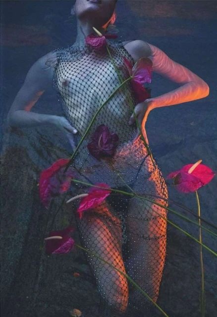 An adult woman with a net-like garment is partially submerged in water, surrounded by red Anthurium flowers in a surreal composition.