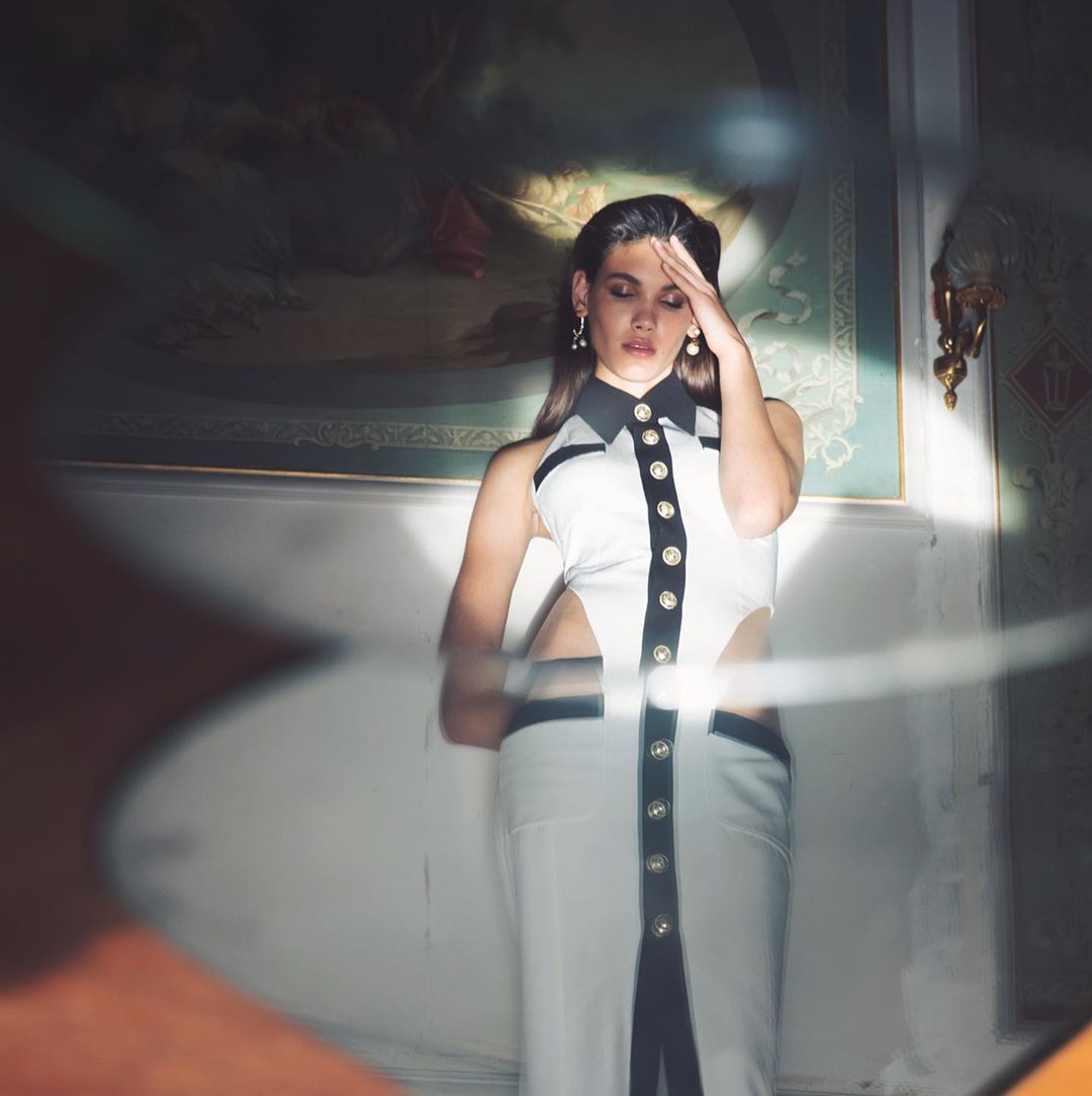 An adult female model posing indoors wearing a buttoned black and white dress with cutouts in a fashion photography style.