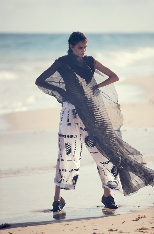 Model wearing patterned white trousers and a sheer black cape standing on a sandy beach by the ocean, photographed by David Bellemere.