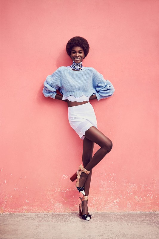 Smiling Black model in a blue sweater and white skirt poses against a pink wall in high heels with ribbons and bows.