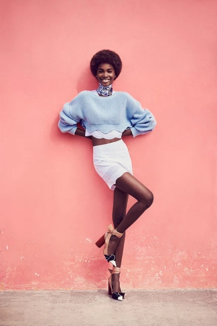 Smiling Black model in a blue sweater and white skirt poses against a pink wall in high heels with ribbons and bows.