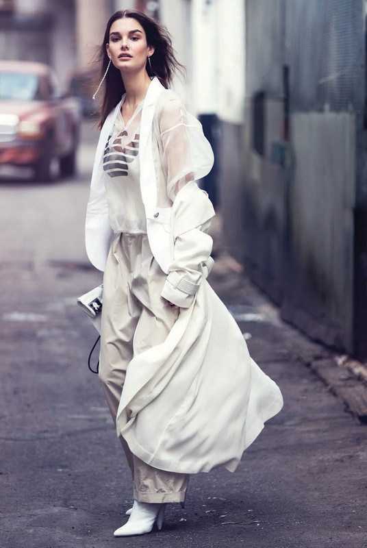 A fashionable adult woman wearing a white outfit walks on a city street with blurred background. She is holding a newspaper.