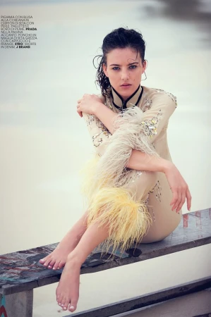 Woman with wet dark hair wearing a feathered beige tunic seated barefoot on a wooden bench outdoors
