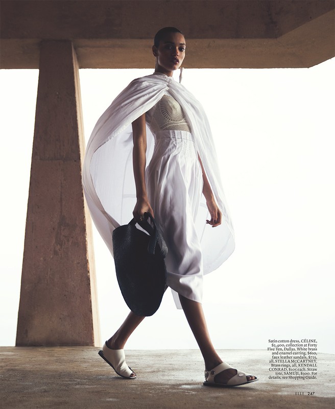Fashion editorial shot of a woman in a white dress and cape walking in an outdoor setting with a dark bag and open-toed shoes.