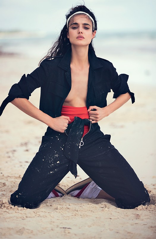 Model kneeling on sandy beach wearing a black utility jumpsuit over a red top and white headband