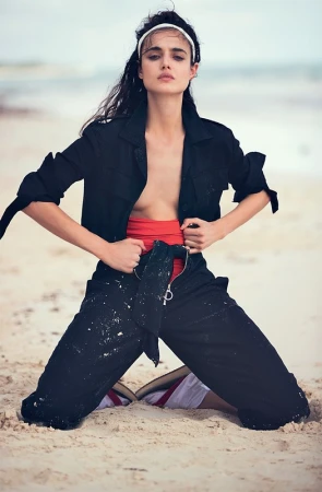 Model kneeling on sandy beach wearing a black utility jumpsuit over a red top and white headband