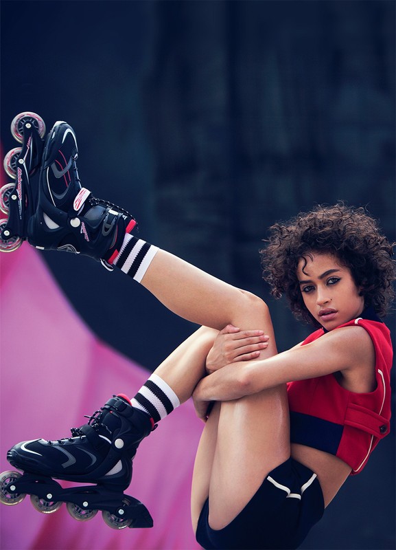 Young woman in athletic wear and roller skates poses with legs raised against a pink and dark blue urban backdrop, looking at the camera.