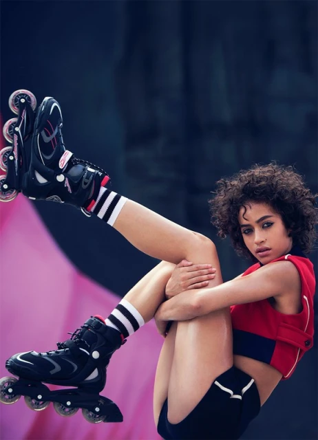 Young woman in athletic wear and roller skates poses with legs raised against a pink and dark blue urban backdrop, looking at the camera.
