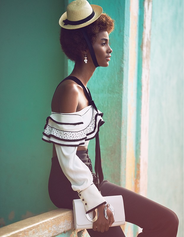 Fashionable young woman with straw hat, wearing a white ruffled top and dark pants, posing against an aqua-colored wall.