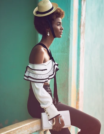 Fashionable young woman with straw hat, wearing a white ruffled top and dark pants, posing against an aqua-colored wall.