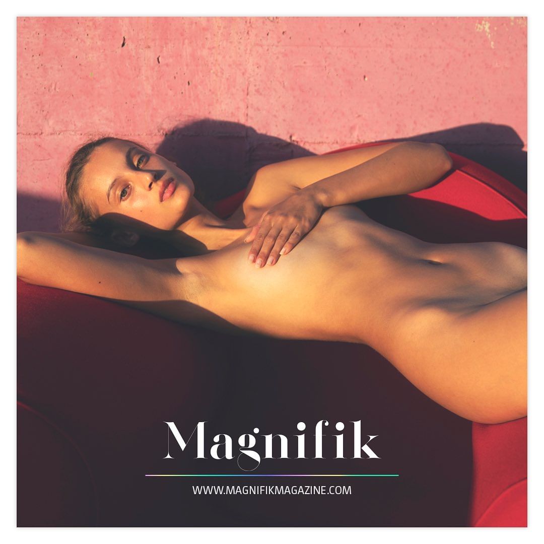 Nude woman poses on a red surface against a pink wall, lit by bright light and shadows. Editorial fashion and beauty image.