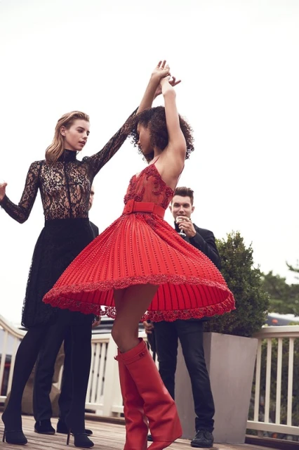 Two women in black and red dresses pose and dance outside with a man in a suit holding a wine glass in the background.