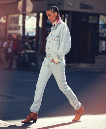 Model with braided hair wearing light wash denim jacket and jeans walking down city street