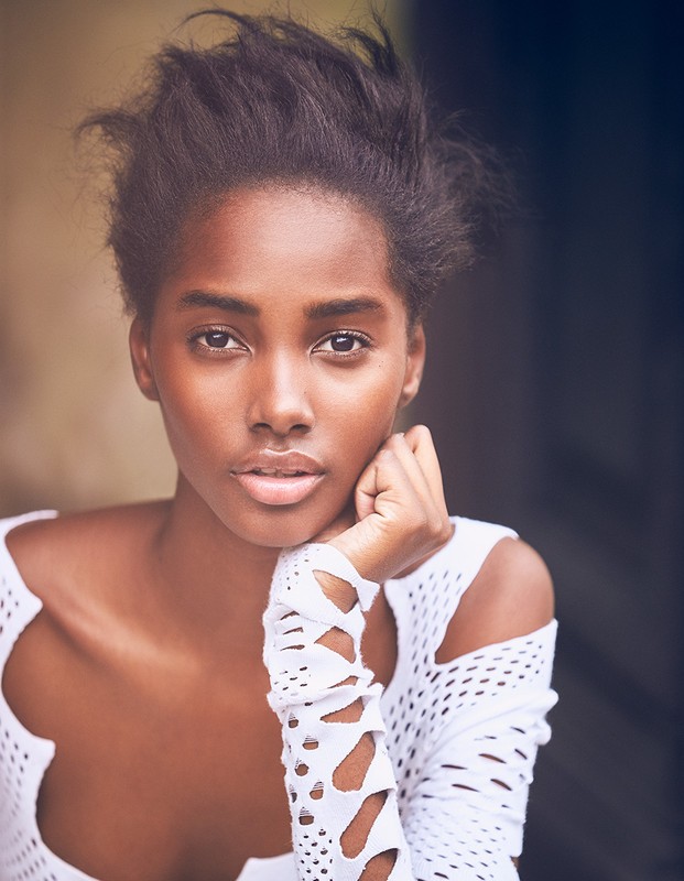 Close-up studio portrait of a Black adult female model wearing a white lattice-patterned top, with her chin resting on her hand and gazing forward.
