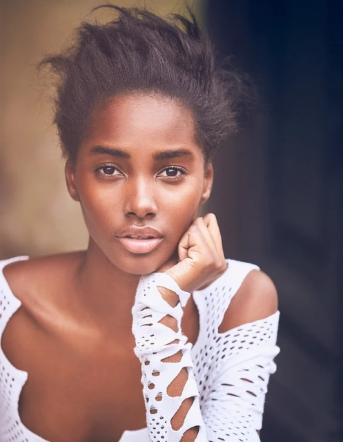 Close-up studio portrait of a Black adult female model wearing a white lattice-patterned top, with her chin resting on her hand and gazing forward.