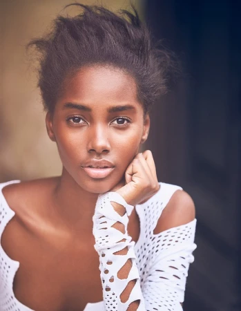 Close-up studio portrait of a Black adult female model wearing a white lattice-patterned top, with her chin resting on her hand and gazing forward.
