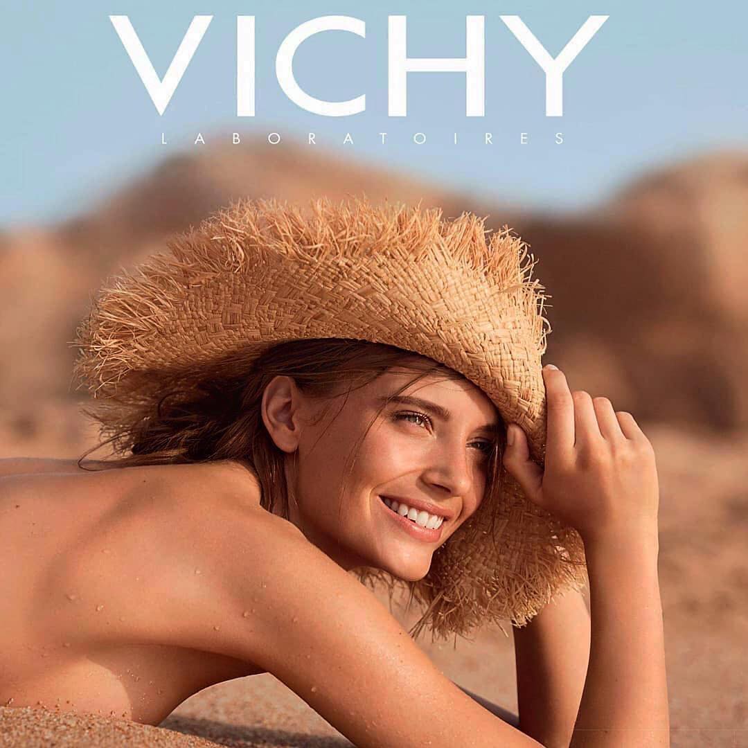 Smiling brunette woman wearing straw hat on sand, Vichy Laboratories advertisement. Sunlit outdoor beauty and skincare product promotion.